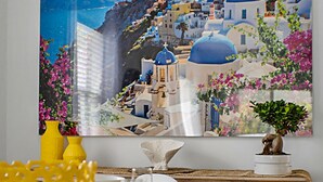 Un comedor con una mesa dispuesta para dos, un cuadro grande de un pueblo costero y una pequeña planta en maceta sobre la mesa.