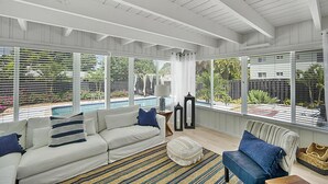 Un salón amplio con techo blanco, ventanas grandes, un sofá blanco, un sillón azul y una mesita de noche con una lámpara.