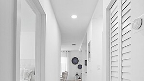 Un estrecho pasillo de paredes blancas con suelo de madera que conduce a un dormitorio con una cama y una silla.