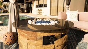 A stone fire pit with a control panel, surrounded by a pool and a patio area with a sofa and a decorative bowl.