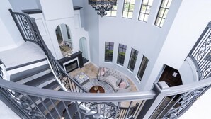 A grand staircase with ornate railings leading to a high-ceilinged room with a fireplace, seating area, and large windows offering views of the outdoors.