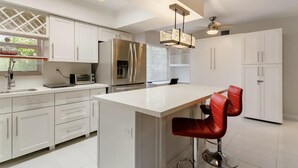 A modern kitchen with white cabinetry, a stainless steel refrigerator, a built-in oven, a coffee maker, and a bar area with red bar stools. - Walk to Fort Lauderdale Beach - Perfect Beach Escape (Fort Lauderdale)