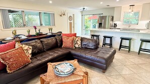 A modern living room with a brown leather sectional sofa, a wooden coffee table, and a dining area with a table set for four. - AVAILABLE NOW!! FERN COTTAGE! FREE beach parking! Weekly/Monthly/Yearly Rentals! (Naples)
