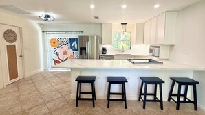 A modern kitchen with a central island, four stools, a refrigerator, a microwave, and a window with blinds. - AVAILABLE NOW!! FERN COTTAGE! FREE beach parking! Weekly/Monthly/Yearly Rentals! (Naples)