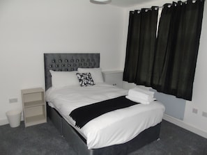 A neatly made bed with white linens and a black throw blanket, a grey upholstered headboard, and black curtains on a window.