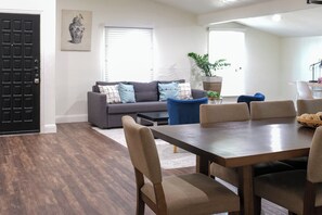 Ein modernes Wohnzimmer mit dunklem Holzboden, einem grauen Sofa mit gemusterten Kissen, einem blauen Sessel, einem Holzesstisch mit beigen Stühlen und einer schwarzen Tür.