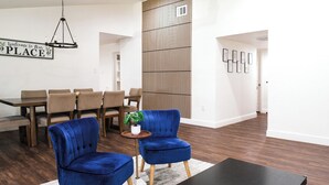 Ein modernes Wohnzimmer mit Essbereich, mit einem schwarzen Couchtisch, zwei blauen Sesseln, einem Holzessttisch mit Stühlen und einem dekorativen Schild an der Wand.