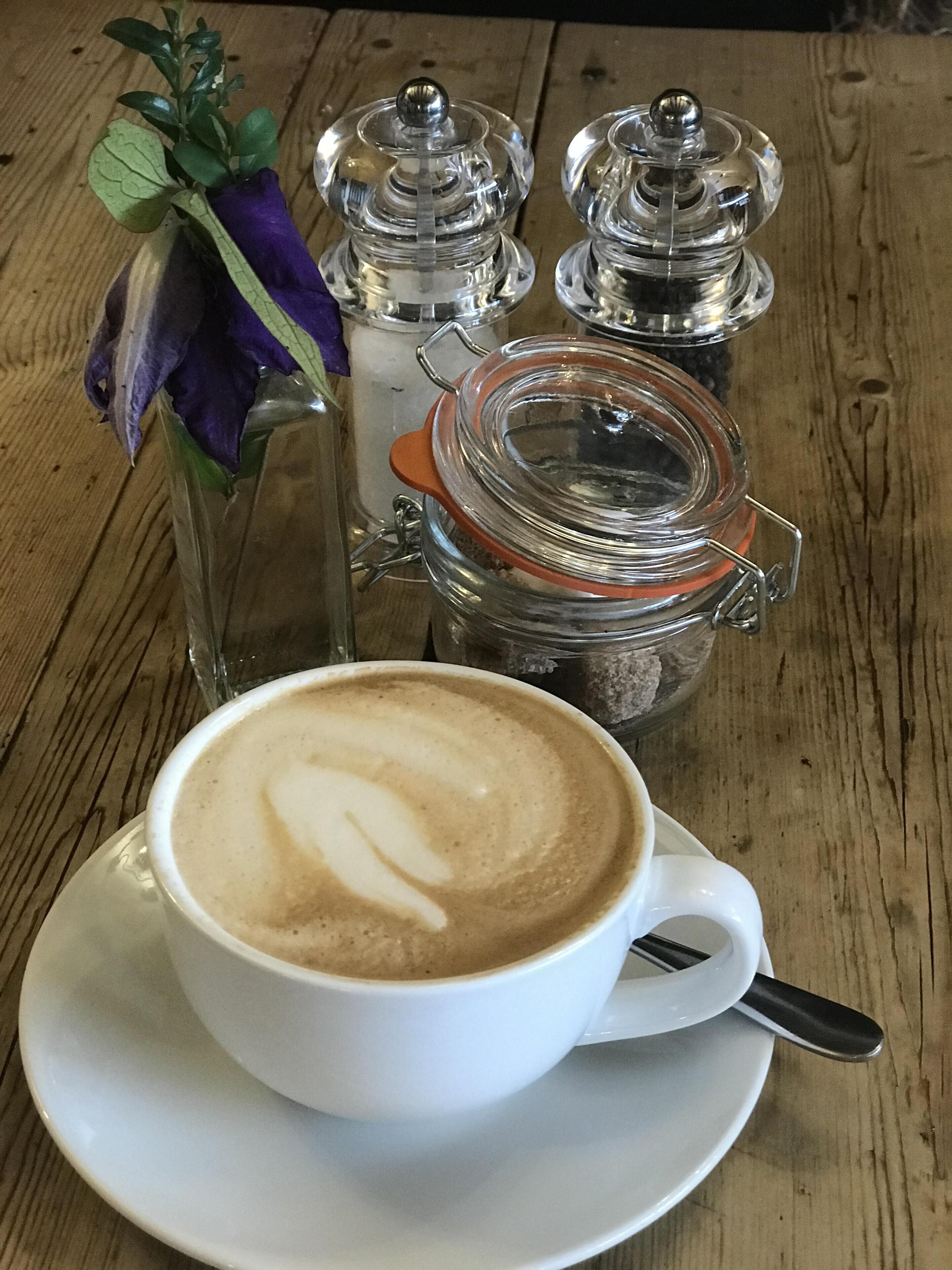 A cup of coffee with latte art on a white saucer, a glass jar with a metal lid, and a glass vase with a purple flower on a wooden table.