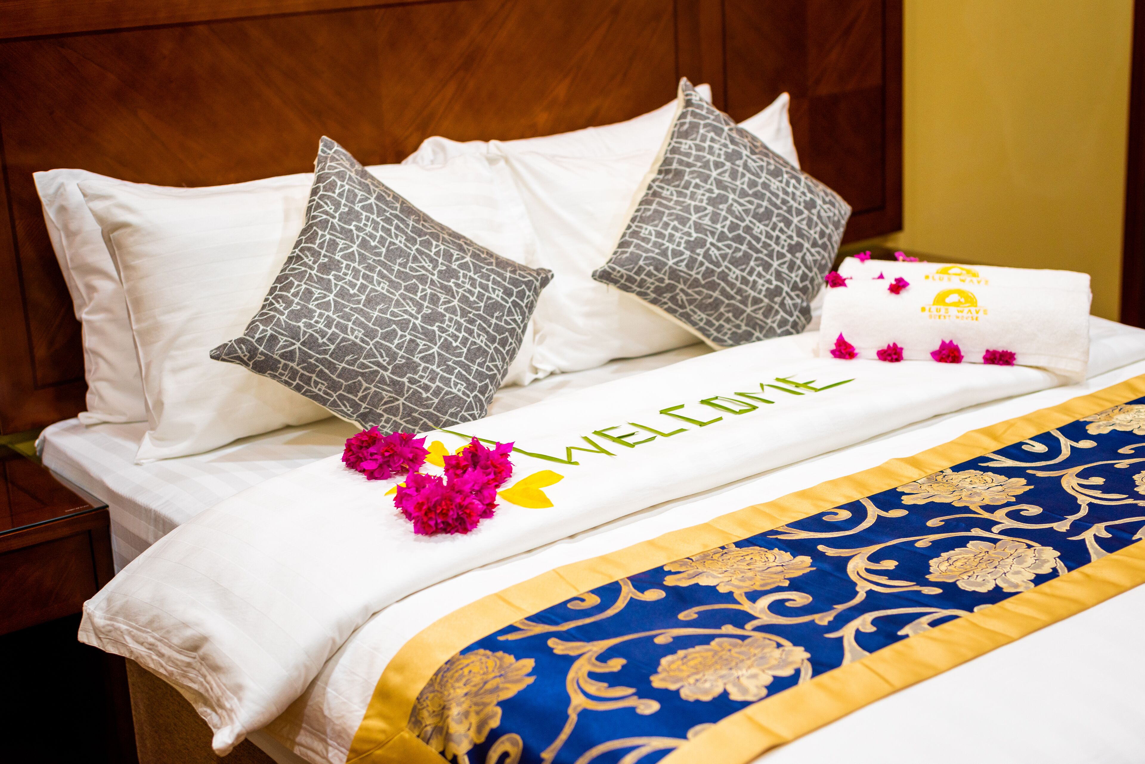 A neatly made bed with a 'WELCOME' sign, pillows, and a decorative blue and gold bedspread.