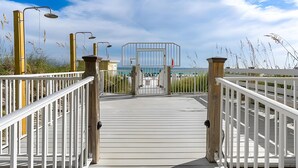 Een houten terras leidt naar een hekpoort, met een helder uitzicht op het strand en de oceaan op de achtergrond.