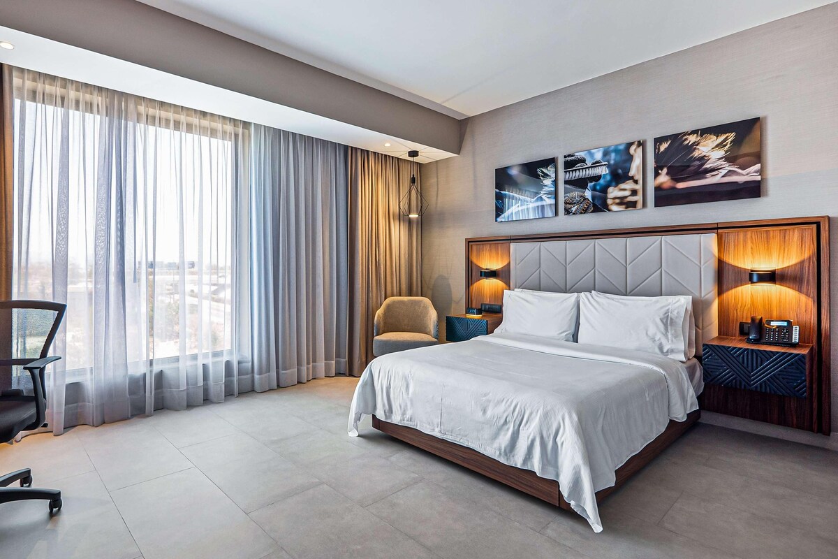 A modern hotel room with a large bed, a desk, a chair, and a view of the city through large windows.