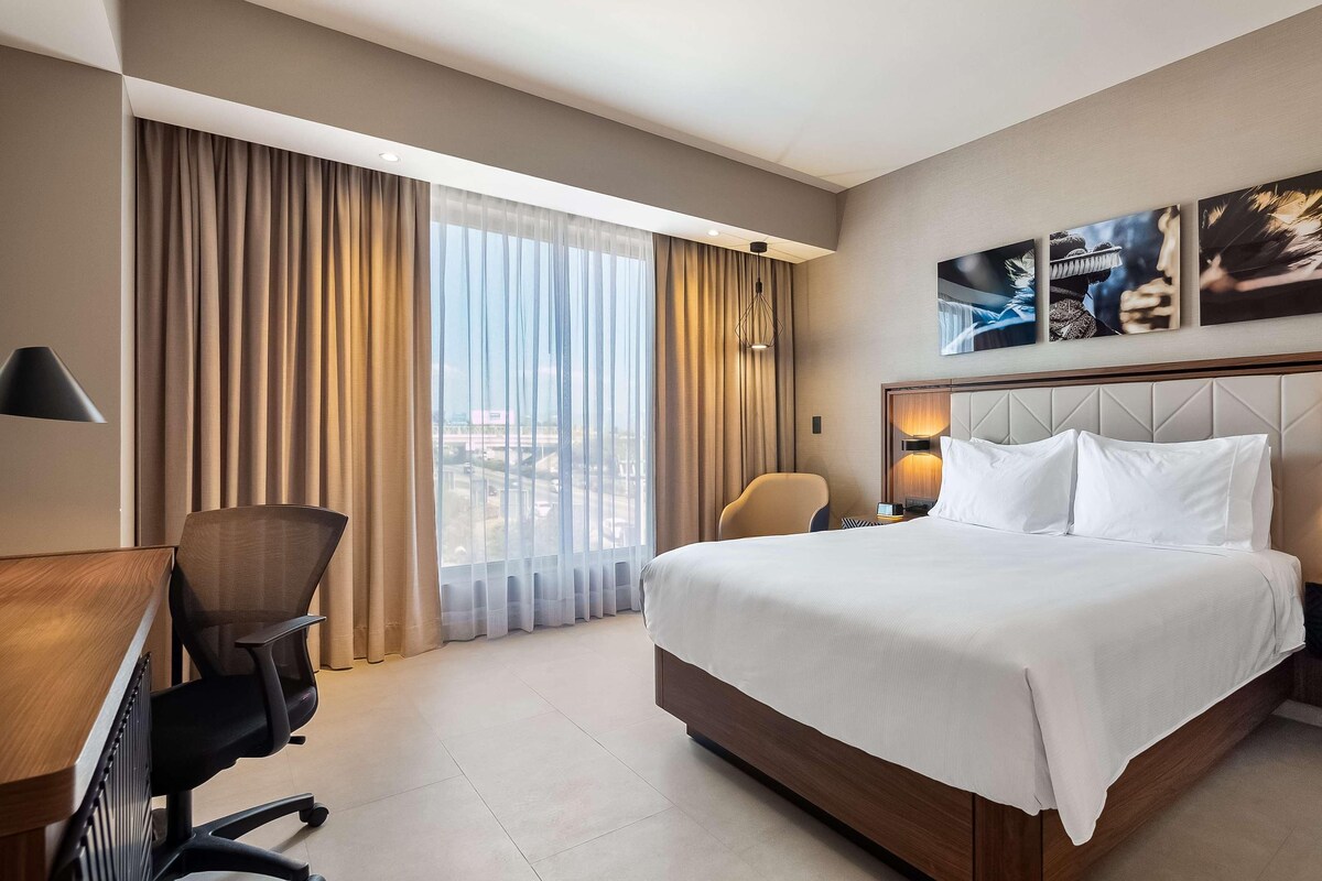 A hotel room with a large bed, a desk, a chair, and a view of the city through large windows.