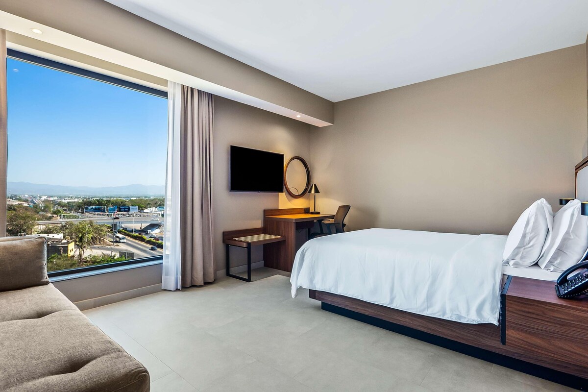 A hotel room with a large window, a bed, a desk, a chair, and a TV.
