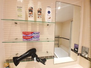 A bathroom with a glass shelf holding toiletries, a black hairdryer, and a mirror. - Livestay 3 Bed Apt on 8th Floor With Amazing Views (London)