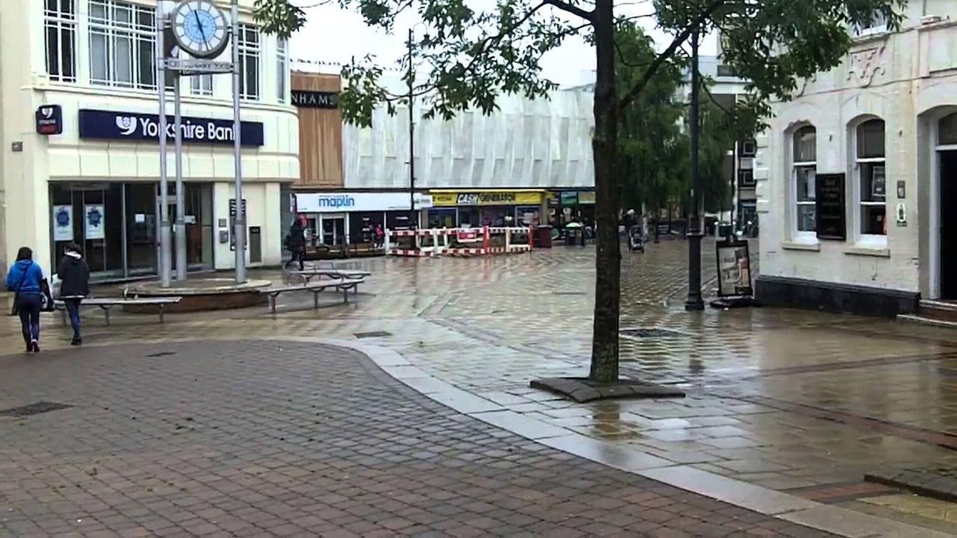 A wet urban square with a clock, a Yorkshire Bank, and a maplin store.