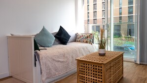 A cozy room with a white sofa, a small wooden table with a plant, and a view of a building through a glass door. - Livestay - 1bed Apt Private Building Free Parking (Luton)