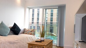 A modern bedroom with a large window, a bed with multiple pillows, a wooden side table, and a television on a stand. - Livestay - 1bed Apt Private Building Free Parking (Luton)