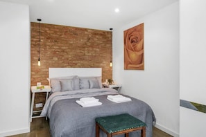 A bedroom with a brick wall, a bed with grey bedding, a green bench, and a painting of a rose.