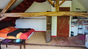 Ein Schlafzimmer mit schräg gedämmter Decke, einem Bett mit roter und orangefarbener Decke, einem Holztisch mit einem Buch und einer roten Tür.