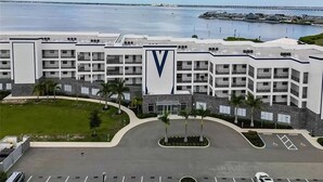 Een meerlaags hotel met een opvallend 'V'-logo, een parkeerplaats en zicht op het water en de jachthaven op de achtergrond.