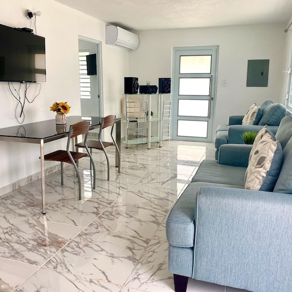 A modern living room with a sofa, a dining area with chairs, a TV mounted on the wall, and an air conditioning unit.