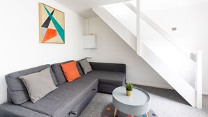 A modern living room with a grey sectional sofa, a small round table with a plant, and a colorful abstract painting on the wall. - Dan Suite by Koya Homes (Barry)