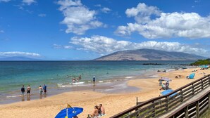 Una playa con un paseo marítimo de madera, gente caminando y nadando, y una montaña al fondo.