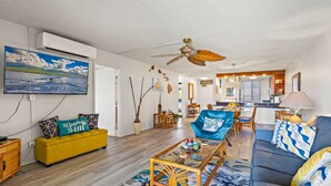 Una sala de estar con un sofá amarillo, un sillón azul, una mesa de centro de vidrio y un ventilador de techo.