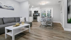 A modern apartment with a grey sofa, a white coffee table, a dining area with a glass table and chairs, a kitchen with stainless steel appliances, and a large window with a view of greenery. - Renovated Home in Hollywood (Hollywood)