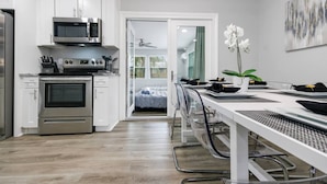 A modern kitchen with white cabinets, a built-in oven, and a microwave. A dining area with a table and chairs is visible, and a bed can be seen through an open door. - Renovated Home in Hollywood (Hollywood)
