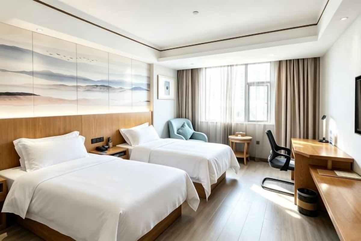 Quarto de hotel com duas camas, uma escrivaninha, uma cadeira e uma janela grande com cortinas.