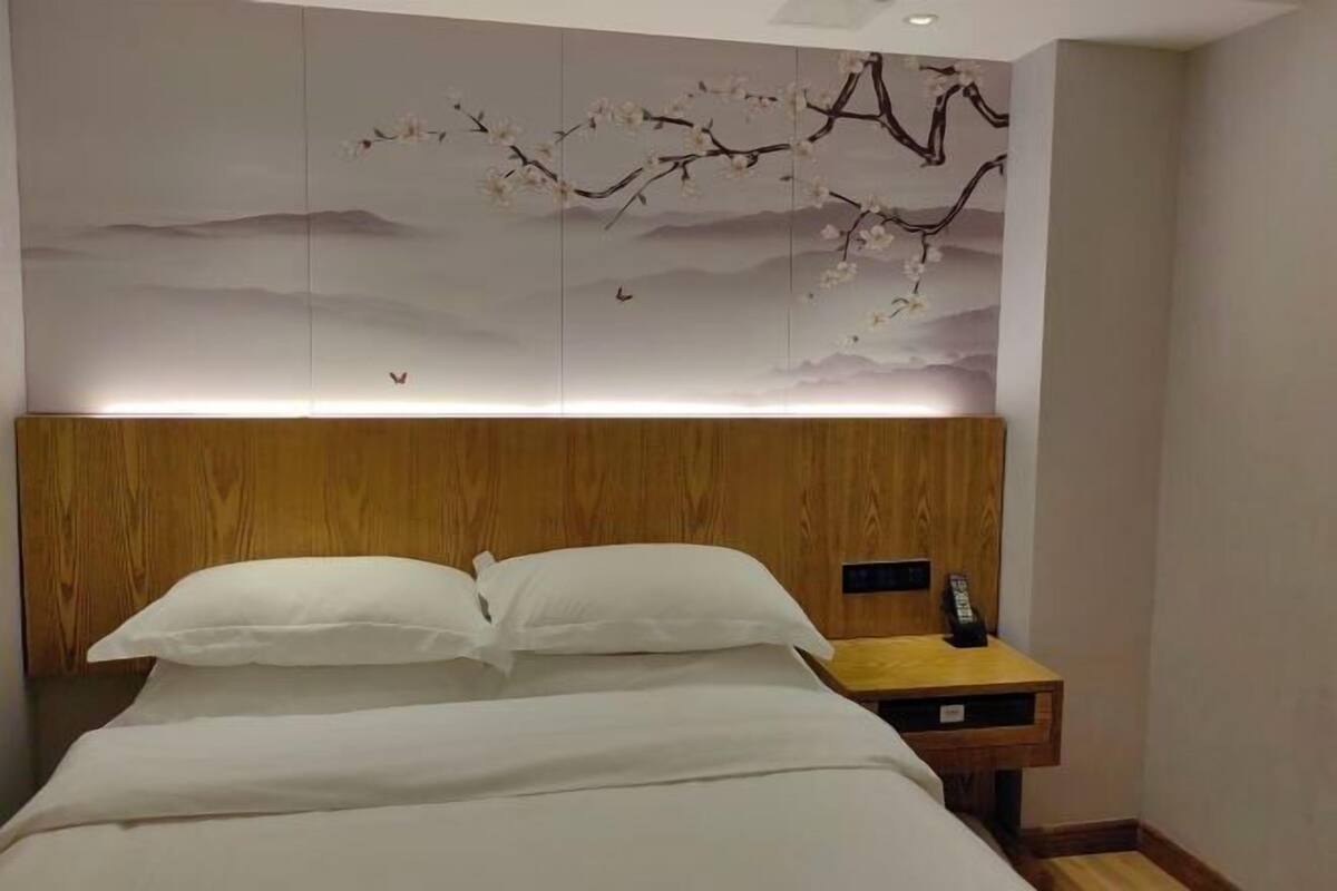 Quarto de hotel com cabeceira de madeira, cama com roupa de cama branca, mesa de cabeceira com controle remoto e um mural de flor de cerejeira na parede.