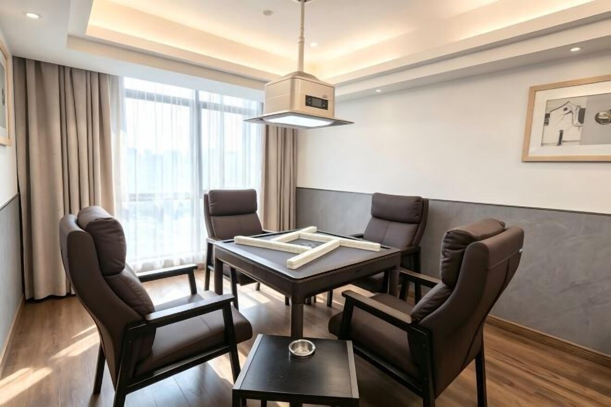 Una habitación de hotel moderna con zona de comedor, una mesa y cuatro sillas, ventanas amplias con cortinas y una lámpara en el techo.