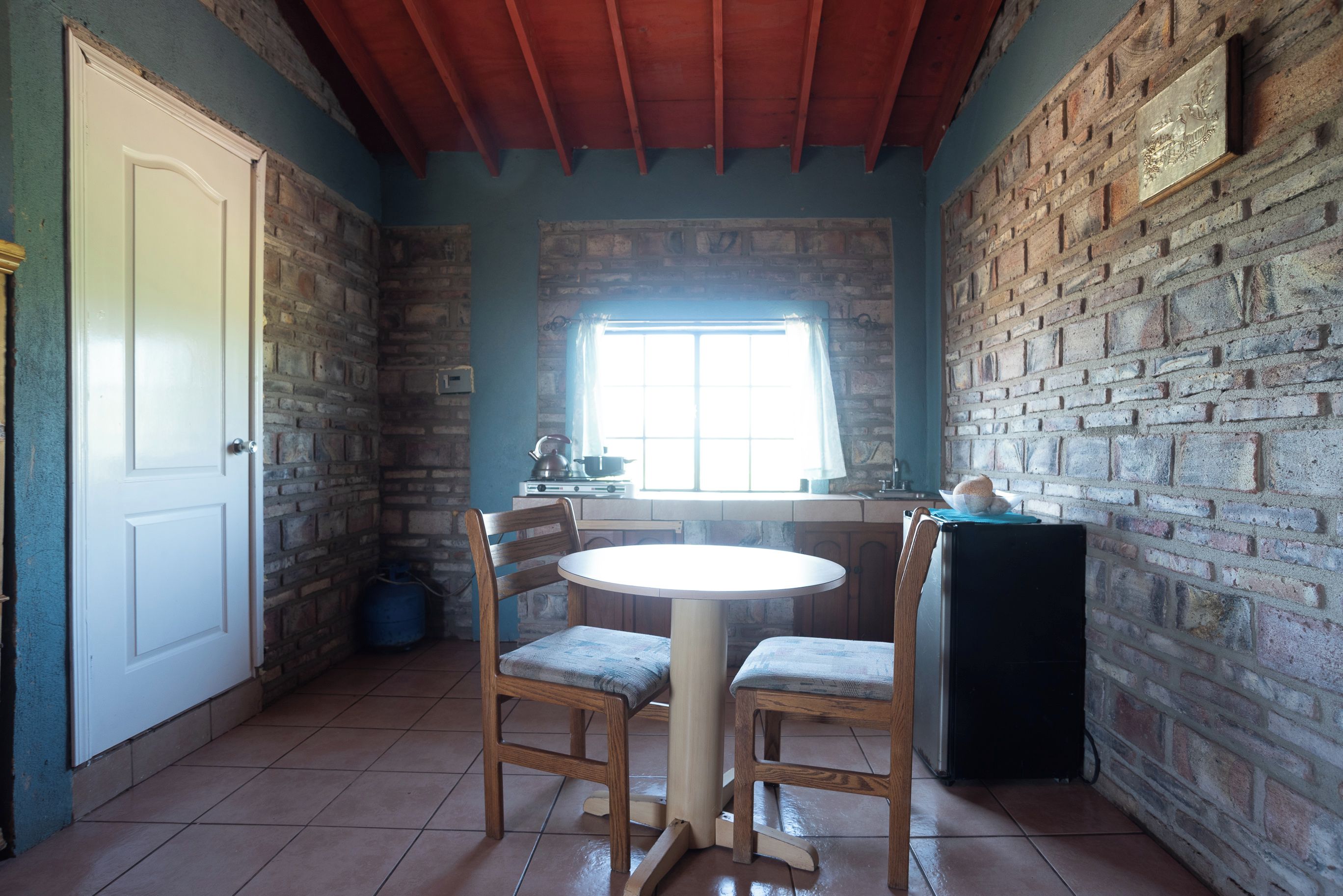 Une pièce avec un mur en briques, une porte blanche, une fenêtre avec des rideaux, une table ronde avec deux chaises et un réfrigérateur.