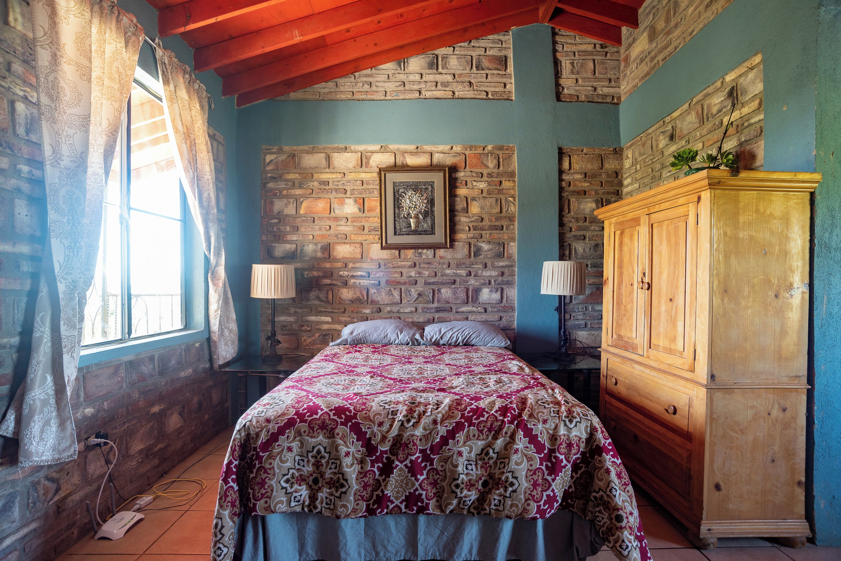 Une chambre avec un mur en briques, un lit en bois, une table de chevet, une armoire et une fenêtre avec des rideaux.