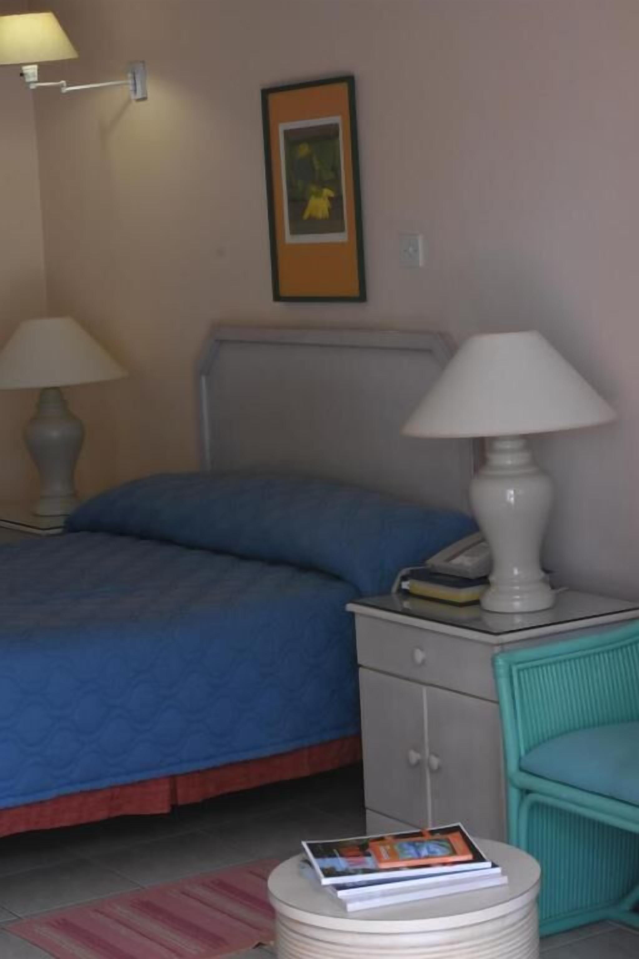 Een slaapkamer met een bed, bedlampjes, een nachtkastje met een lamp, een lijst met een foto aan de muur, en een klein rond tafeltje met een boek.