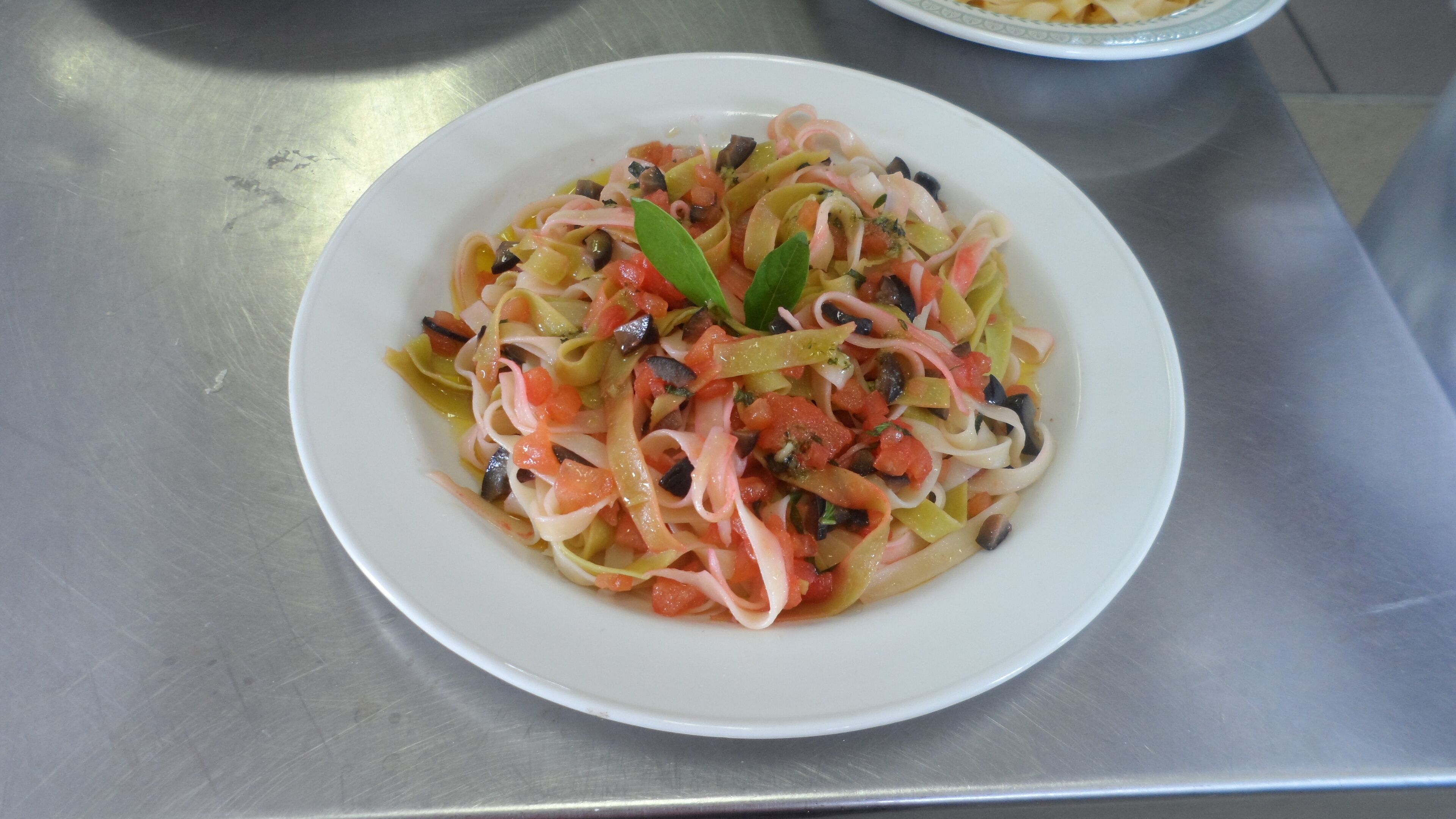 Une assiette de salade de pâtes, agrémentée de légumes et d’herbes aromatiques, posée sur une table en acier inoxydable.