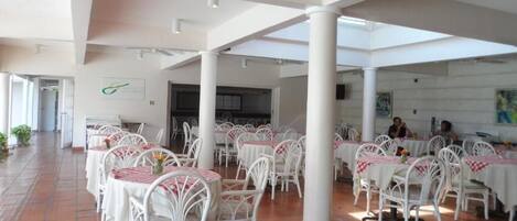 A spacious indoor dining area with white chairs and tables, large pillars, and a tiled floor.