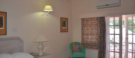 A hotel room with a bed, a nightstand with a lamp, a chair, a window with curtains, and an air conditioning unit.