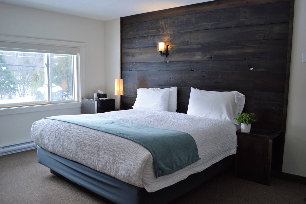A bedroom with a large bed, a nightstand, a wall-mounted light, and a window.