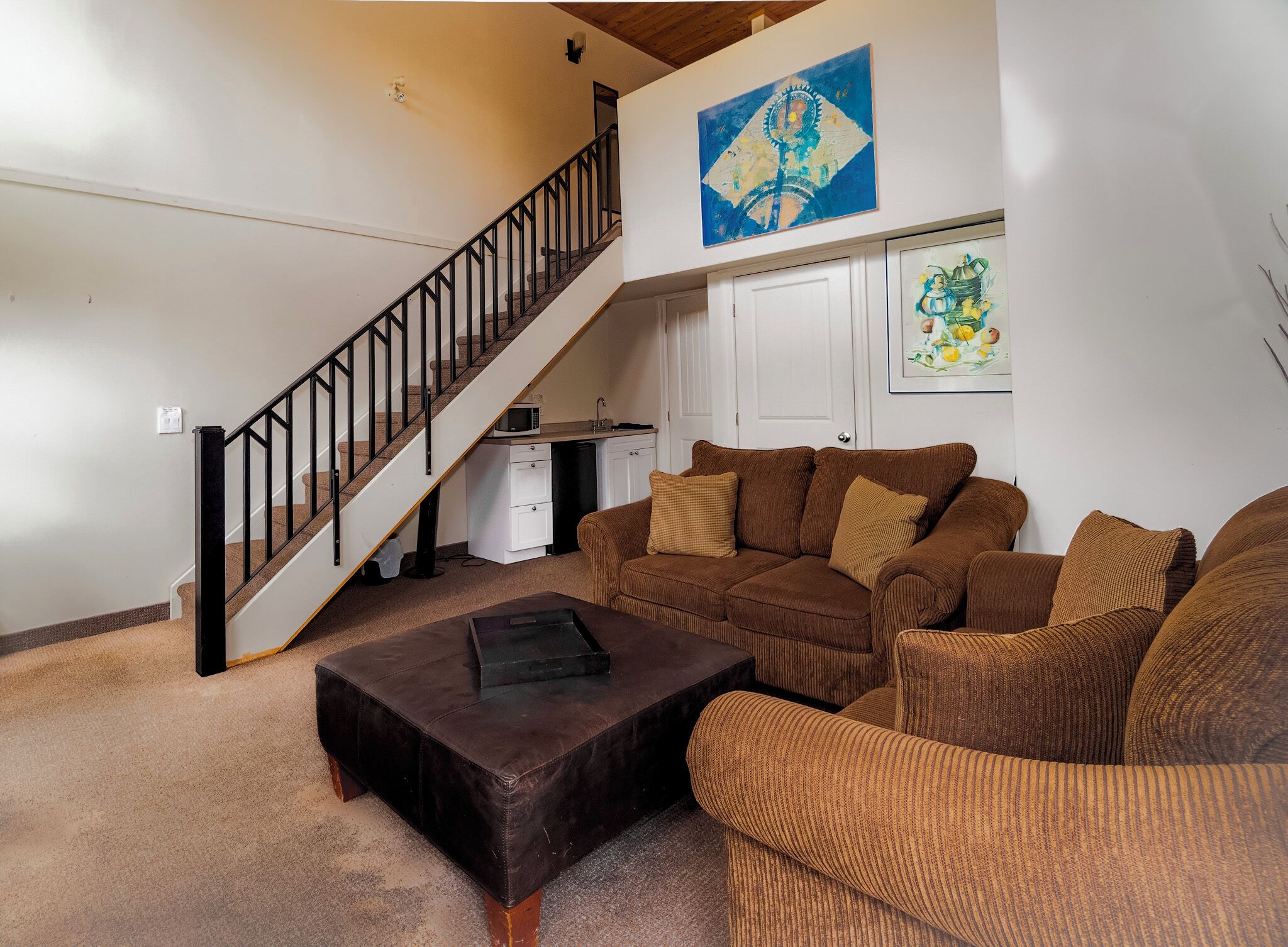 A living room with a brown sofa set, a coffee table, a staircase, a kitchen area, and artwork on the walls.