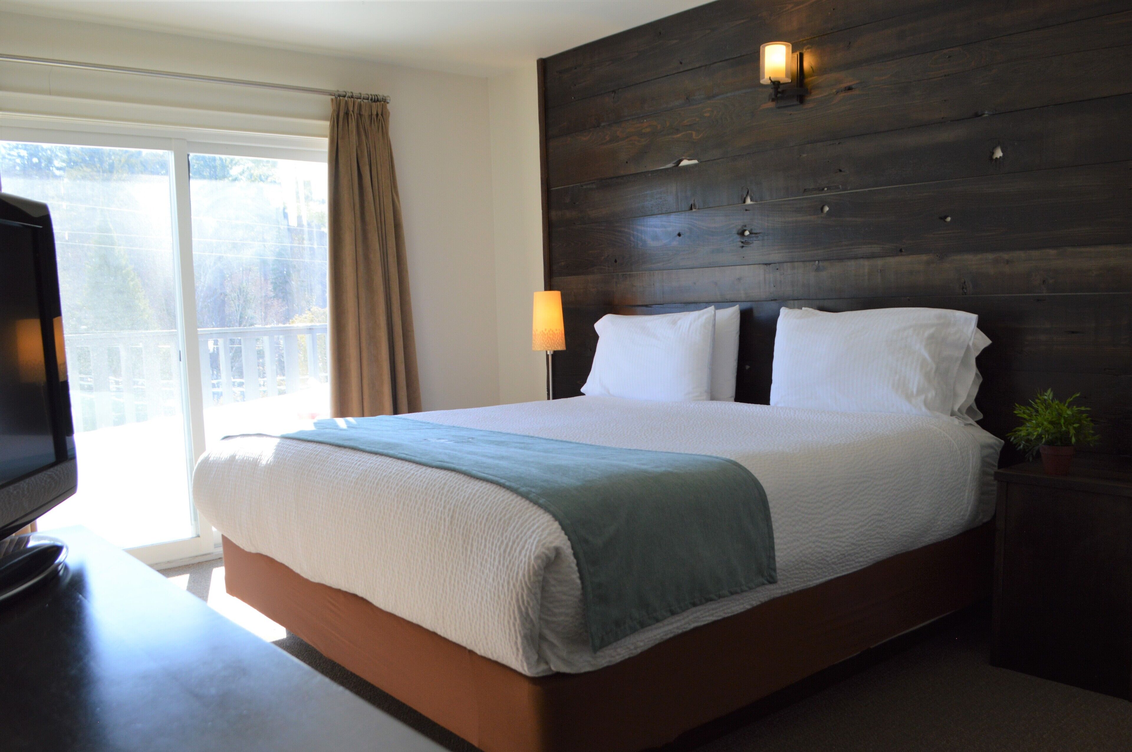 A hotel room with a large bed, a TV on a desk, a window with curtains, and a wooden headboard with wall-mounted lights.