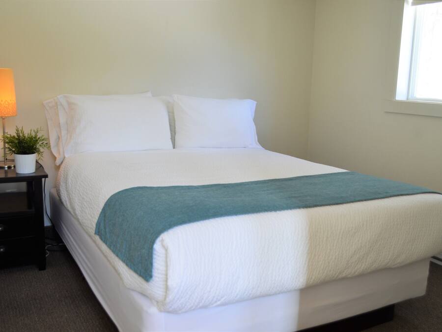 A neatly made bed with white linens and a teal blanket, a bedside lamp, and a small potted plant on a nightstand.