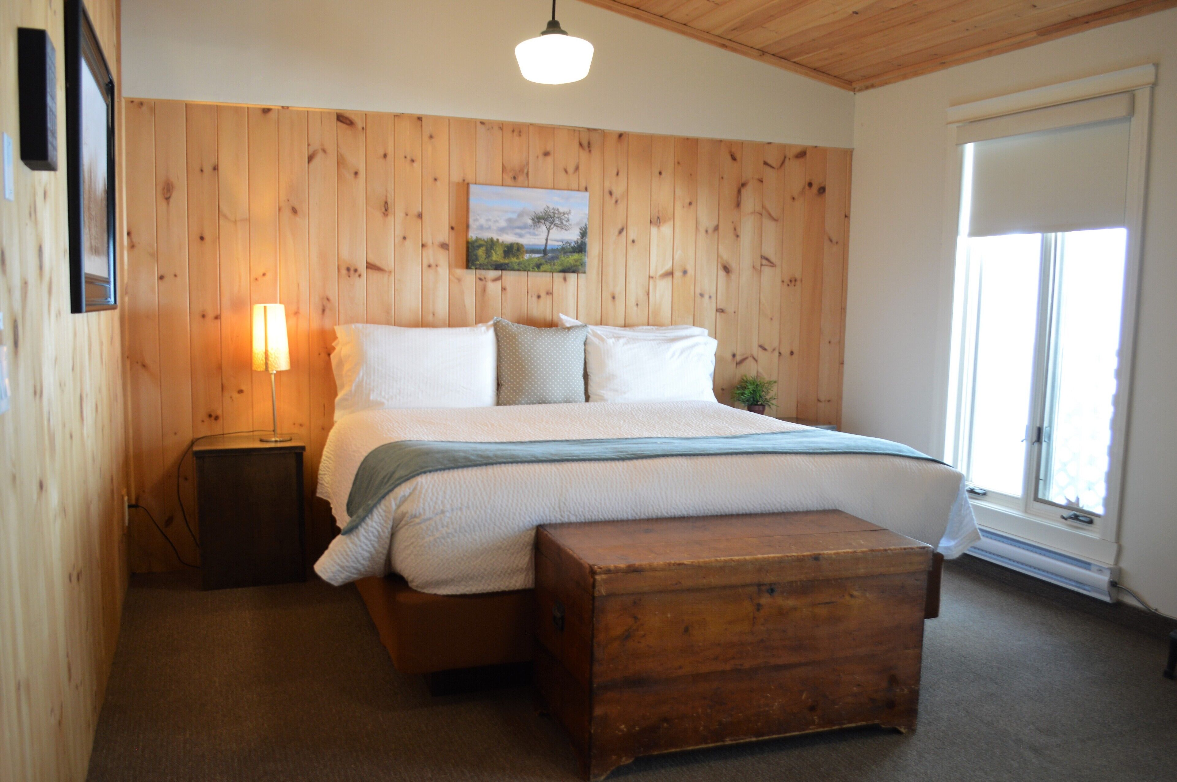 A bedroom with a wooden headboard, a bed with white bedding, a bedside lamp, a wooden chest, and a window with blinds.