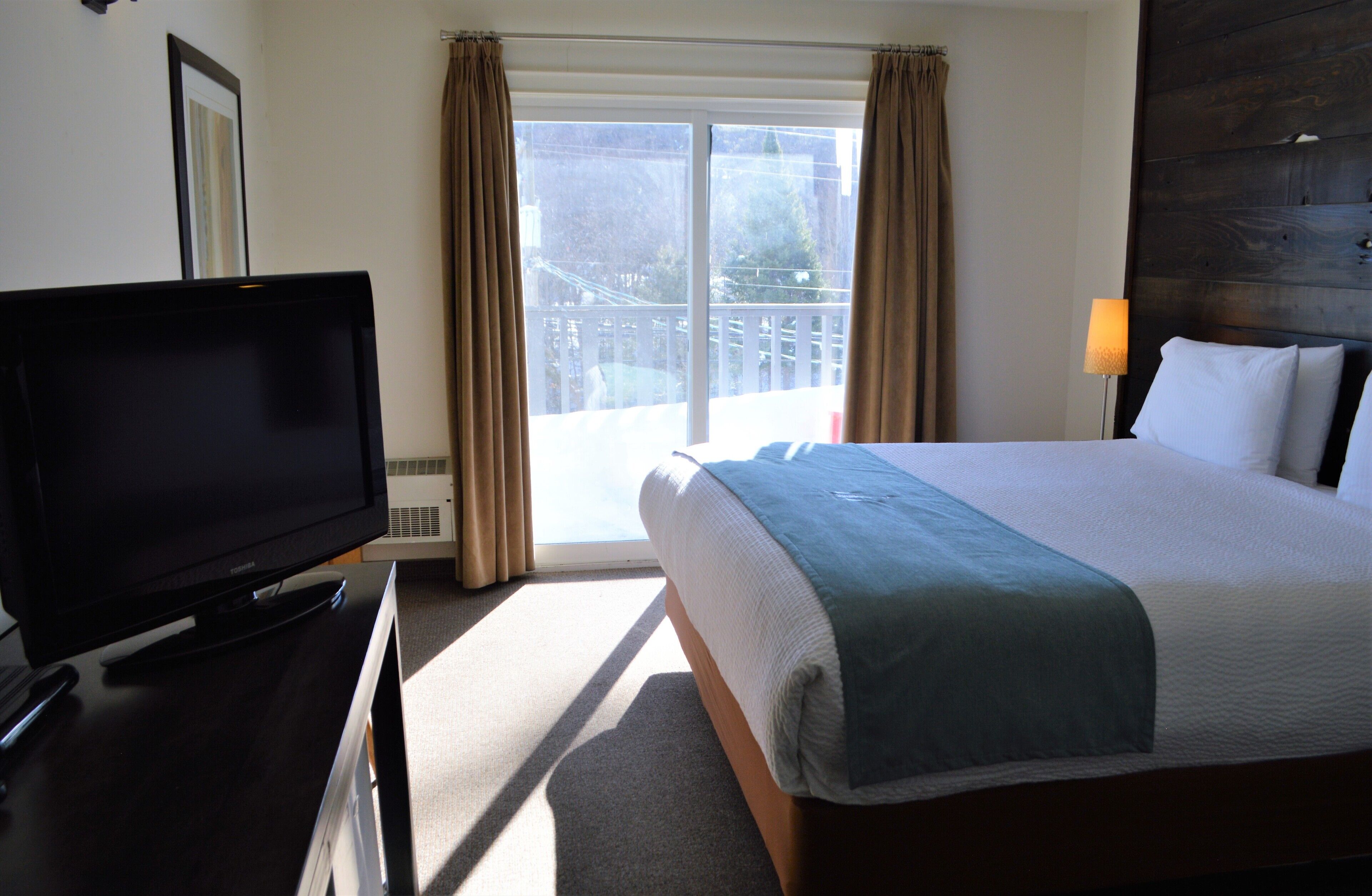 Una habitación de hotel con una cama, televisor, balcón con cortinas y una lámpara.