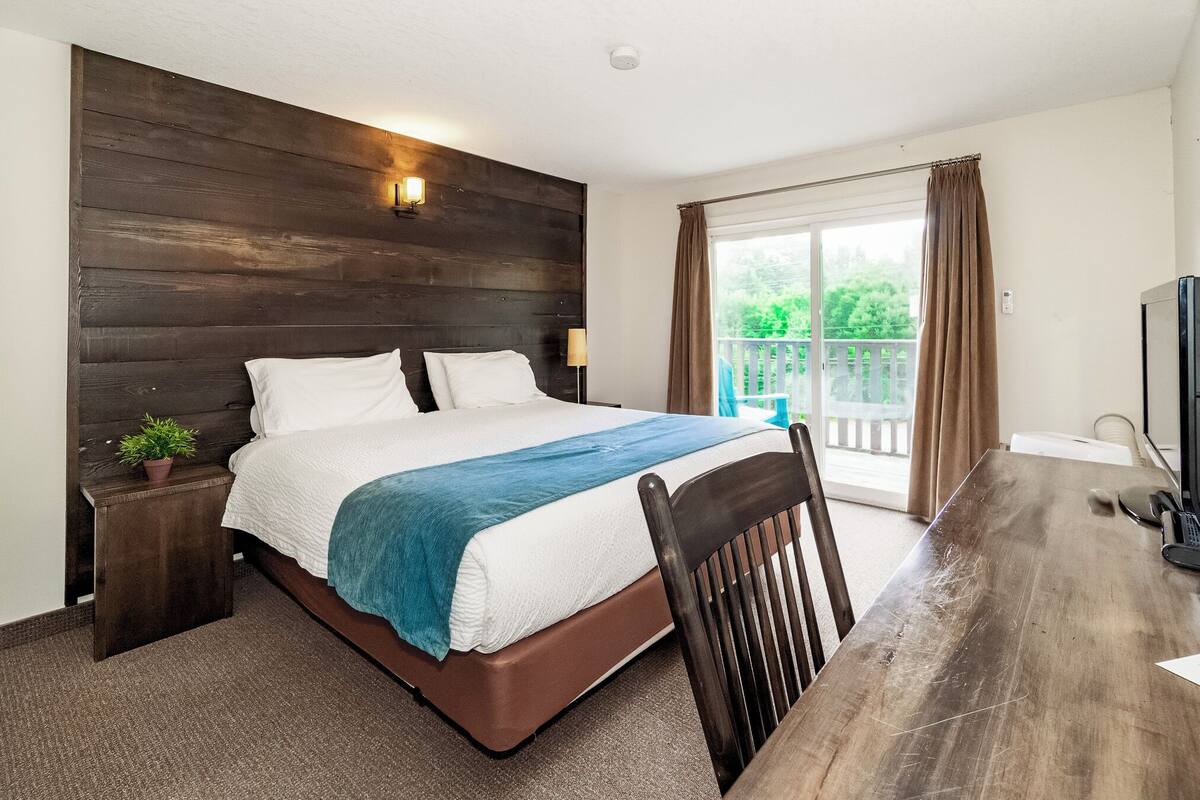 A bedroom with a wooden headboard, a bed with white and blue bedding, a small bedside table with a plant, a wooden desk, and a television.