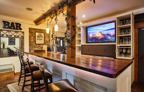 Un espace bar doté d’un comptoir en bois, de tabourets de bar, d’un téléviseur diffusant des images de paysages et d’étagères remplies de verres.