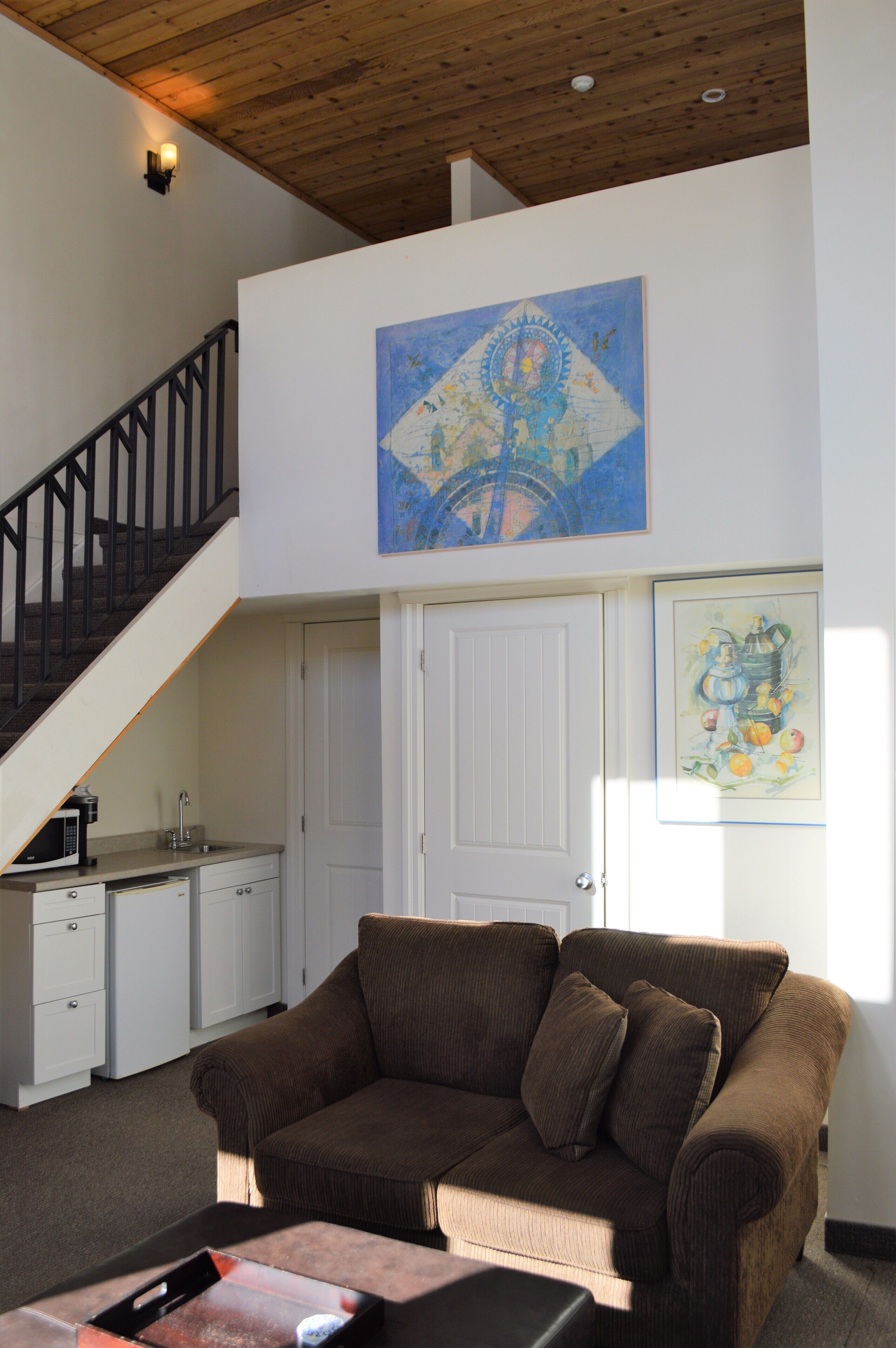 A living room with a brown sofa, a staircase, a kitchen area, and a painting on the wall.
