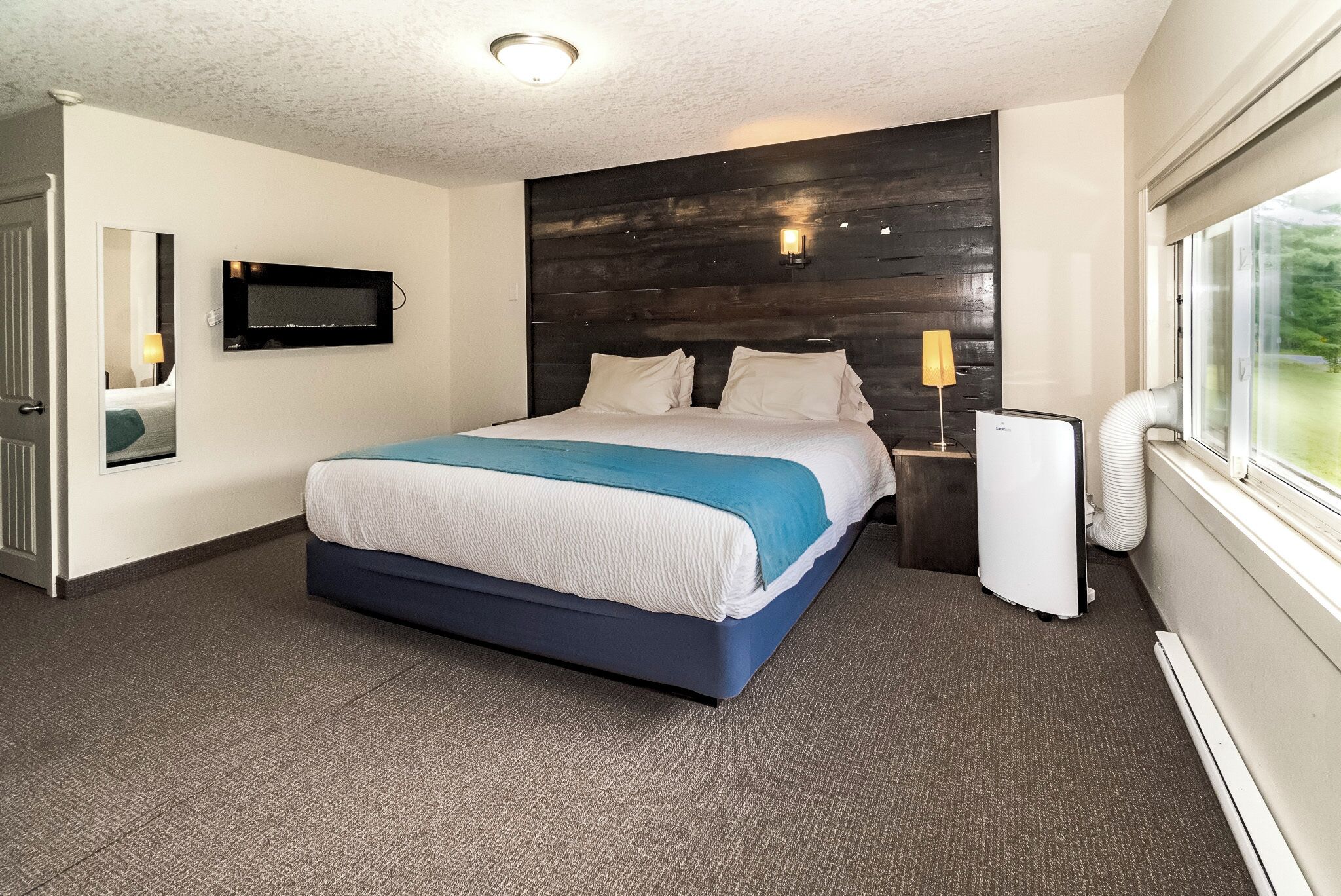 Een moderne hotelkamer met een groot bed, een flatscreen televisie aan de wand, een nachtkastje met een lamp, en een raam met uitzicht op groen.