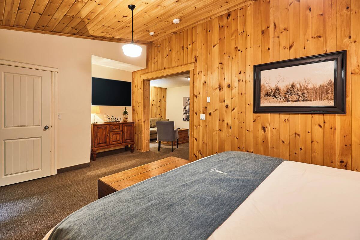 A room with wooden paneling, a bed, a bench, a desk, and a framed picture on the wall.